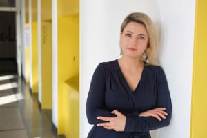 Na zdjęciu dr hab. Barbara Jankowiak, prof. UAM, blondynka w ciemnej sukience przy białej kolumnie.