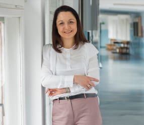 Dr Joanna Dziekońska stoi na korytarzu uczelni, ubrana w białą koszulę i różowe spodnie, oparta o ścianę.