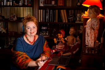 Na zdjęciu prof. dr hab. Dorota Żołądź-Strzelczyk przy biurku na tle półek z książkami i kolekcji lalek.