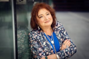 Na zdjęciu prof. Mirosława Nowak-Dziemanowicz, ubrana w kwiecisty żakiet, opiera się o szklaną ścianę.